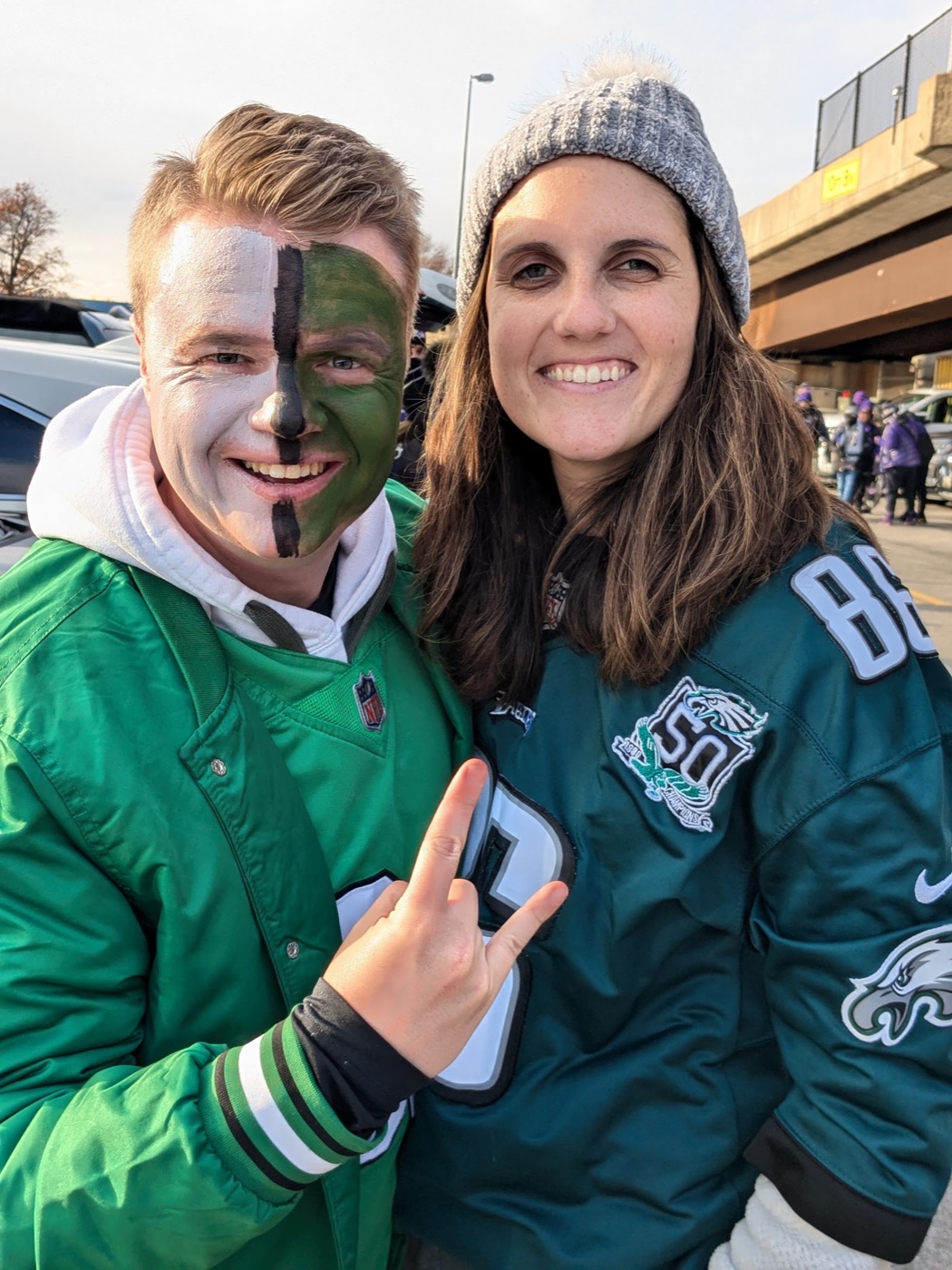Alex Louderback in Eagles face paint and a kelly green jacket at a tailgate, with his wife in a number 88 jersey